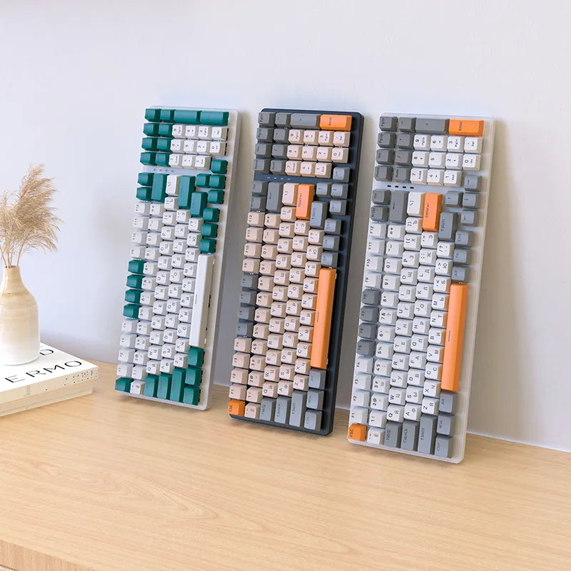 Three colorful mechanical keyboards on a wooden desk: teal, gray, and white with orange accents.